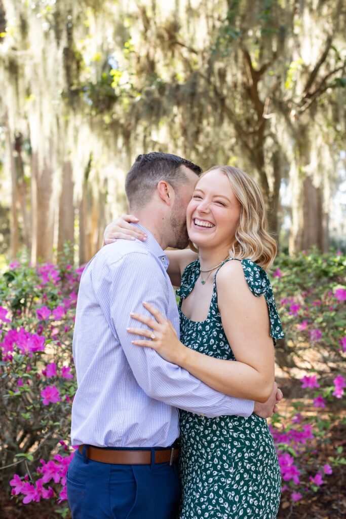 Guy kisses girls cheek making her laugh in front of purple azaleas for their engagement photos at Kraft Azalea Garden in Orlando, Florida