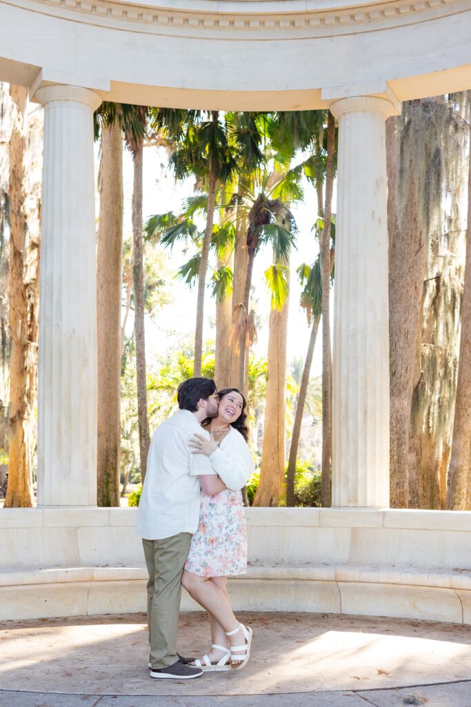 Guy kisses girl's cheek under the columns for their engagement photos at Kraft Azalea Garden in Winter Park, Florida