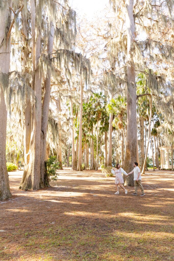 Couple holds hands and runs under live oak trees for their engagement photos at Kraft Azalea Garden in Winter Park, Florida