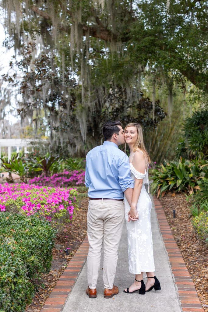 Couple holds hands and guy kisses girls cheek in front of purple flowers for their engagement photos at Cypress Grove Park in Orlando, Florida