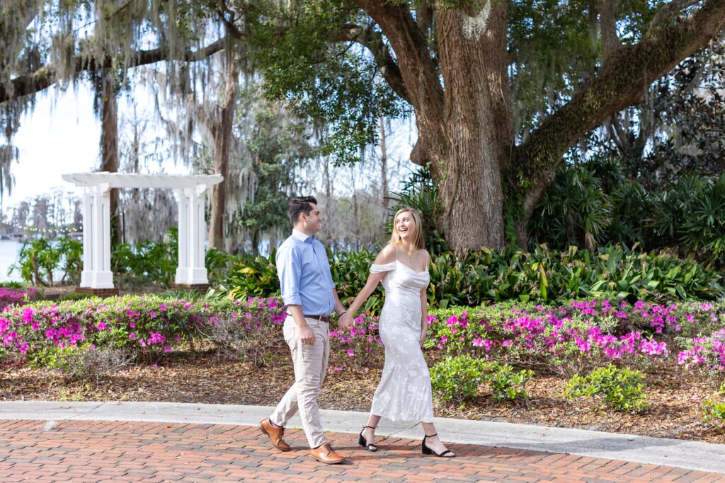 Couple holds hands and walks in front of purple flowers for their engagement photos at Cypress Grove Park in Orlando, Florida