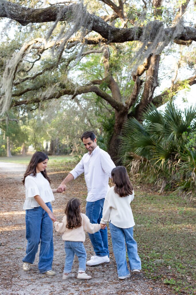 Dad holds hands with daughters under spanish moss tree for their family photos at Greenwood Lakes Park in Lake Mary, Florida with Orlando Family Photgrapher Amy Britton Photography