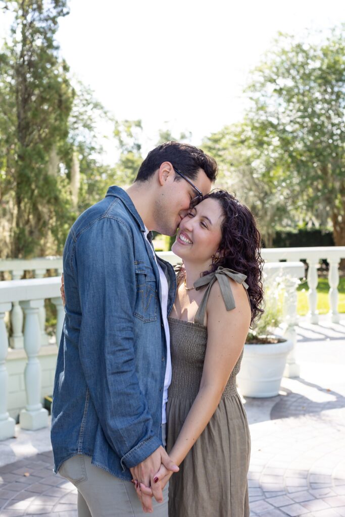 Guy kisses girl's cheek and they hold hands in front of rose garden fountain for their engagement photos at Leu Gardens in Orlando, Florida