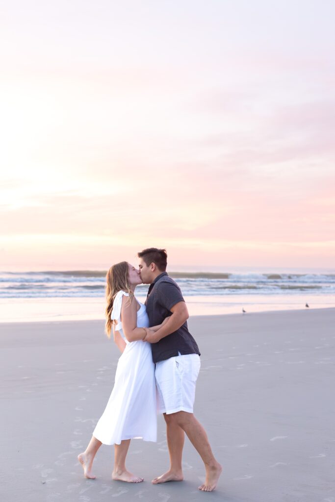 Couple kisses on the beach at sunrise for their engagement photos at New Smyrna Beach, Florida