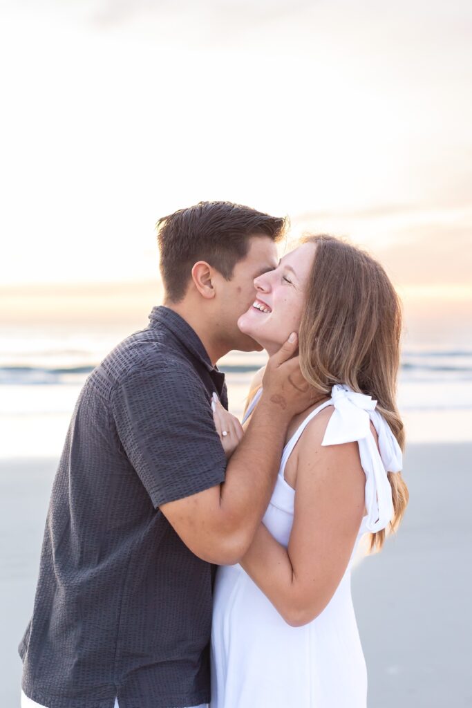 Guy kisses girl's cheek on the beach at sunrise for their engagement photos at New Smyrna Beach, Florida