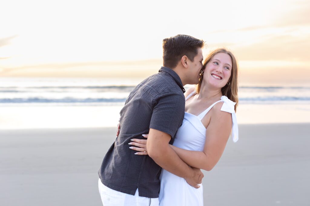 Guy kisses girl's cheek on the beach at sunrise for their engagement photos at New Smyrna Beach, Florida
