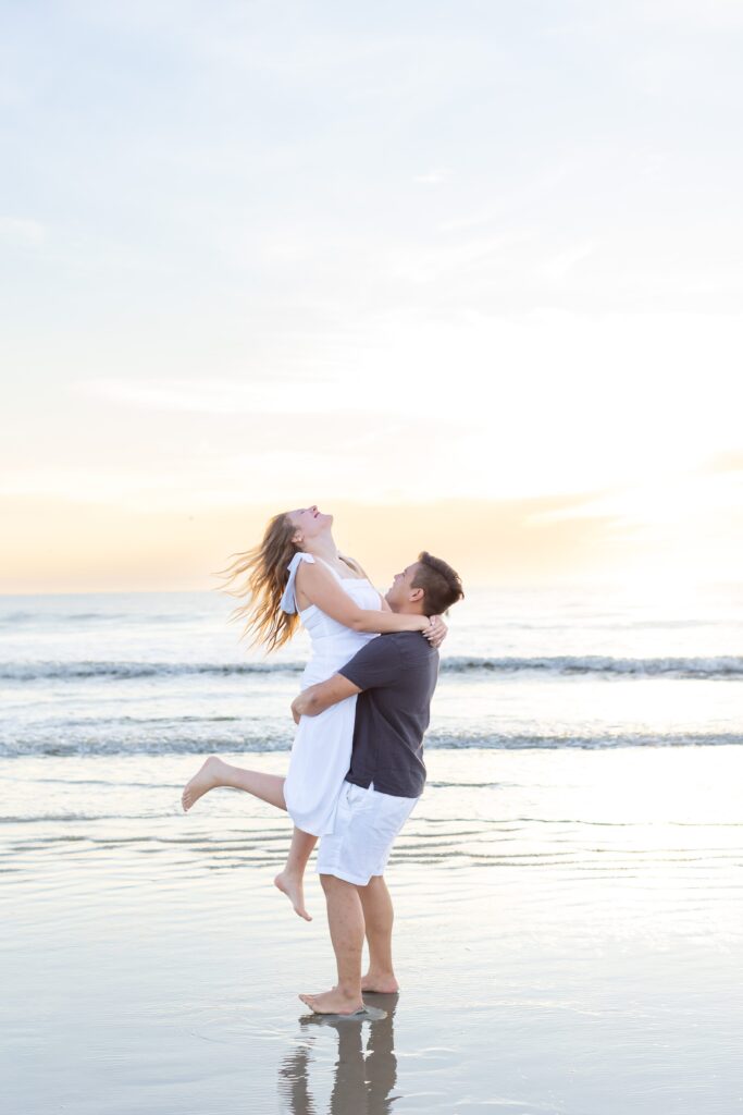 Guy lifts girl and kisses her on the beach at sunrise for their engagement photos at New Smyrna Beach, Florida
