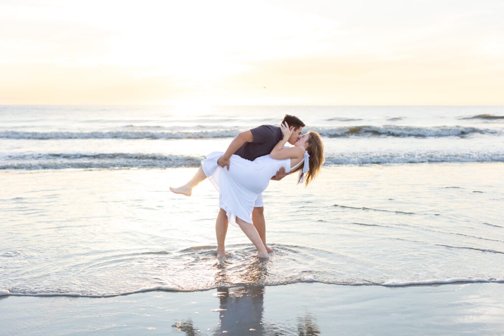 Guy dips girl and kisses her on the beach at sunrise for their engagement photos at New Smyrna Beach, Florida