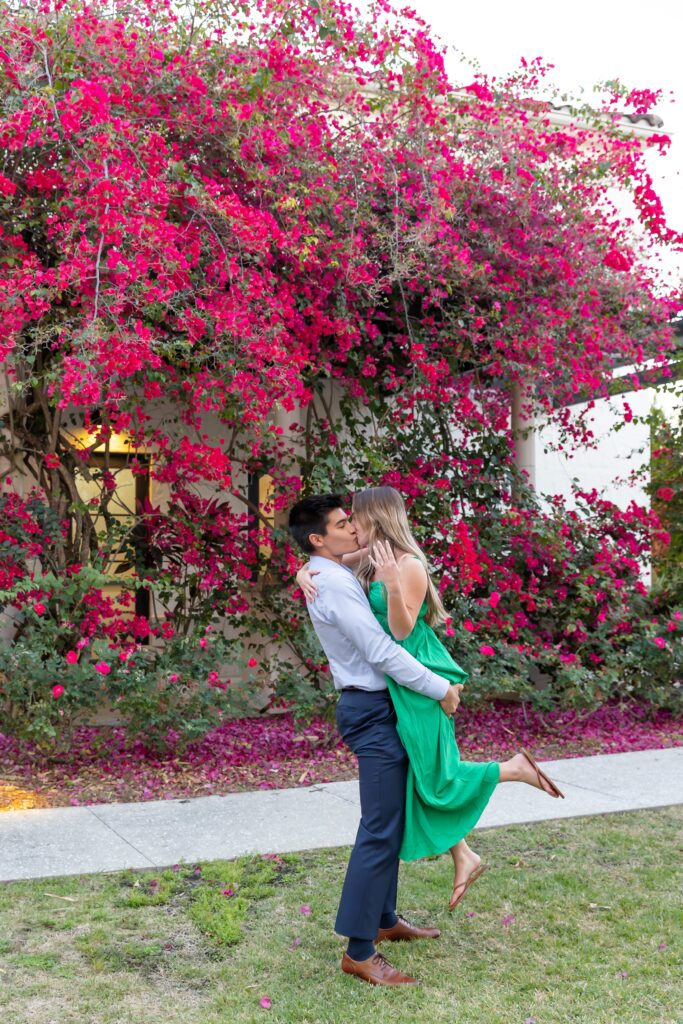 Guy lifts and kisses girl in front of pink flower wall for their engagement photos at Alfond Inn Winter Park, Florida