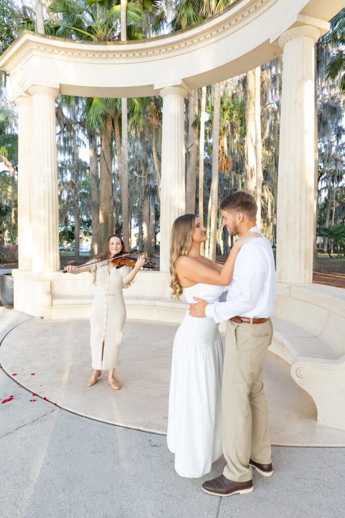 Guy proposes to girl with violinist and rose petal heart under the columns at Kraft Azalea Garden in Winter Park, Florida