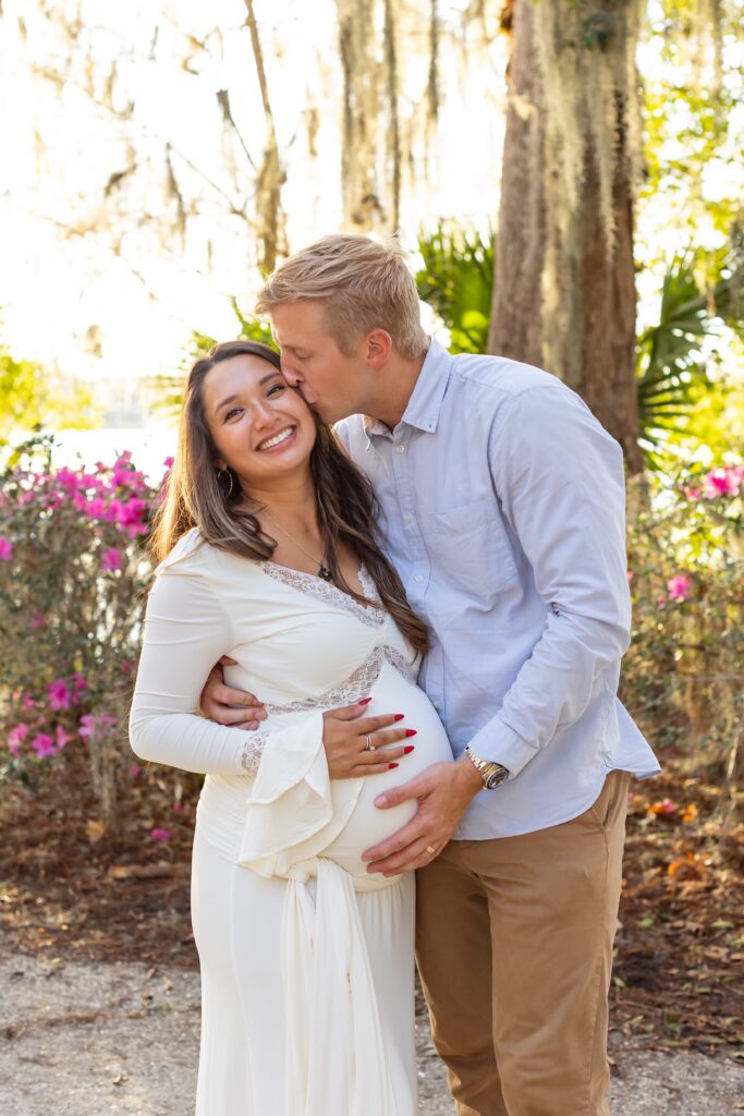 Couple holds wife's belly and husband gives wife kiss for their Maternity Photography Orlando session at Kraft Azalea Garden in Winter Park, Florida