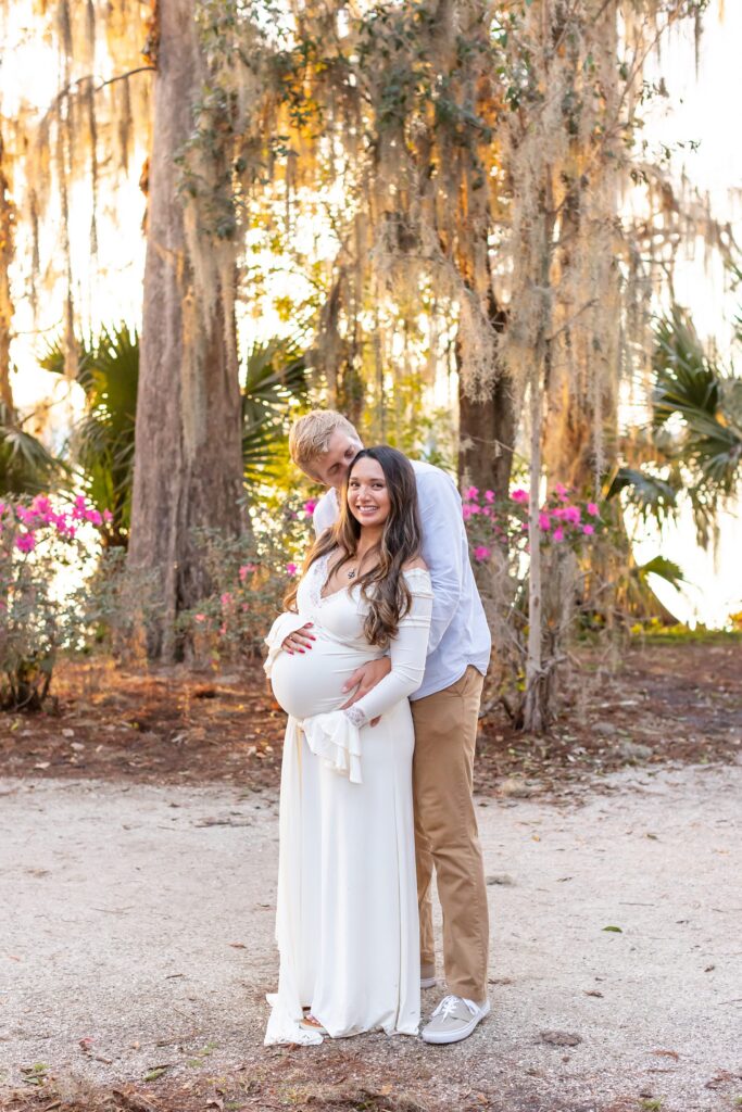 Couple holds wife's belly and husband gives wife kiss for their Maternity Photography Orlando session at Kraft Azalea Garden in Winter Park, Florida