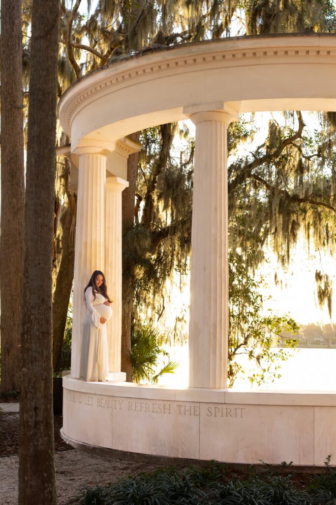 Soon to be mom holds belly and poses on top of columns at sunset at Kraft Azalea Garden in Winter Park Florida for her Maternity Photography Orlando session