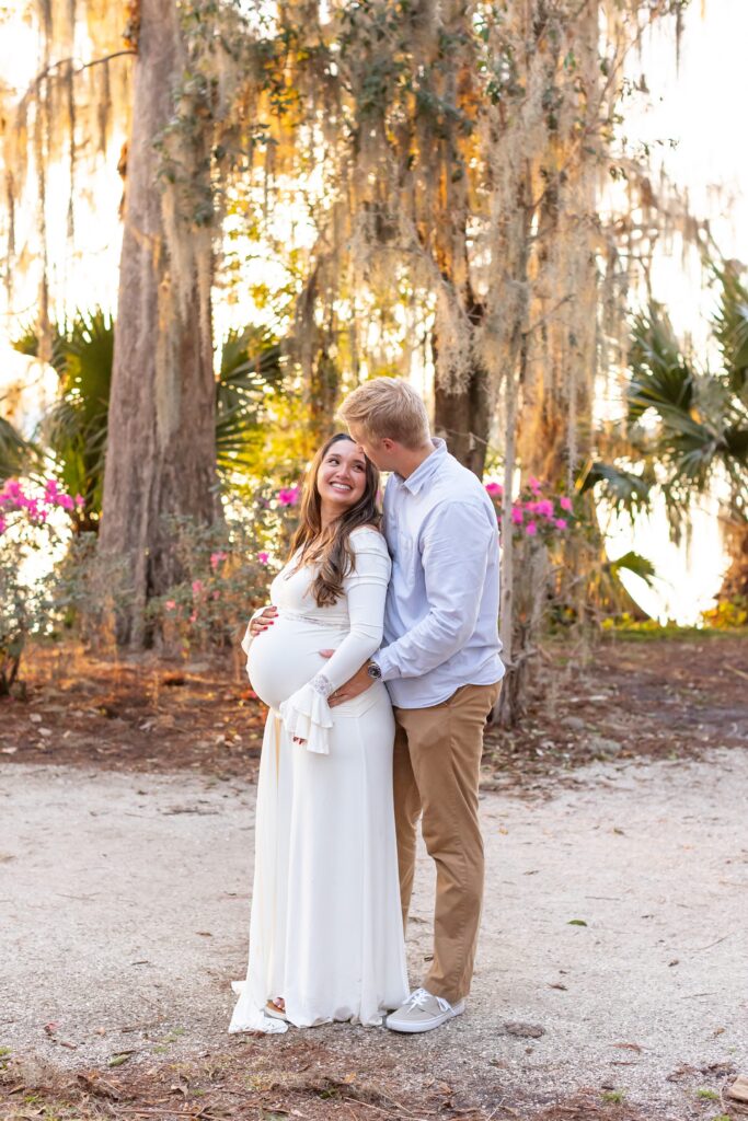Couple holds wife's belly and husband gives wife kiss for their Maternity Photography Orlando session at Kraft Azalea Garden in Winter Park, Florida
